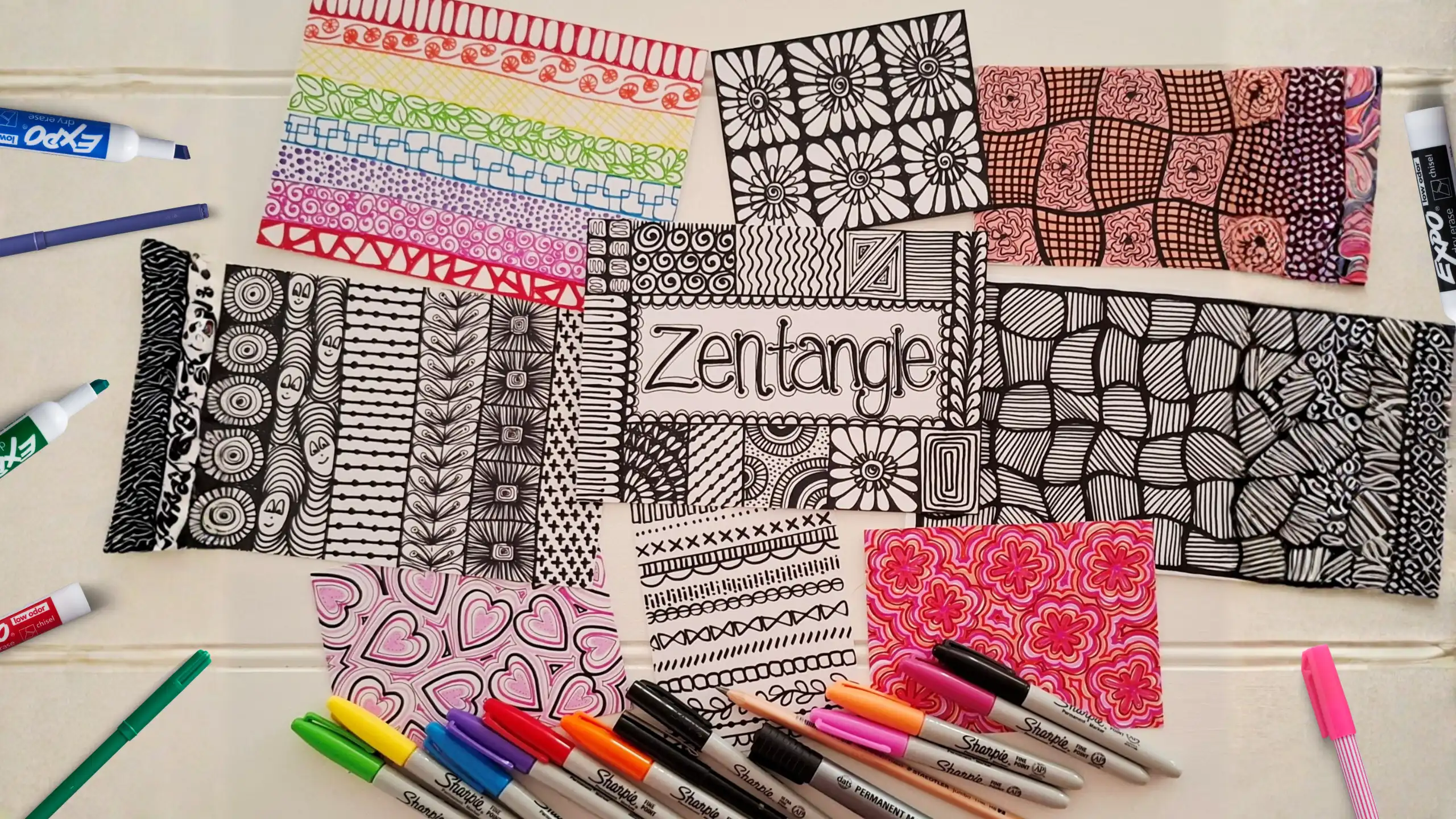 Zentangle-inspired artwork samples displayed on a table, featuring intricate black-and-white patterns, colorful doodle designs, floral motifs, hearts, geometric shapes.